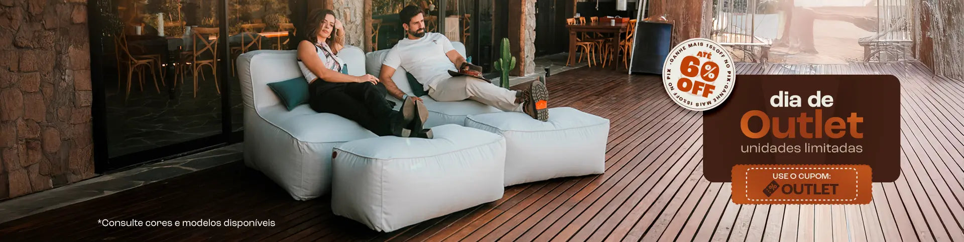 Casal sentado em um sofá cinza em uma sala de estar aconchegante. Eles estão sorrindo e segurando canecas laranja. No fundo, uma estante com livros e objetos decorativos. No topo da imagem, o texto 'Mês dos NAMORADOS' com um coração laranja, e à direita, um banner laranja com o texto 'Site até 35% OFF +30 DIAS PARA TESTAR'. O casal está usando meias coloridas e parecem estar relaxados e felizes.